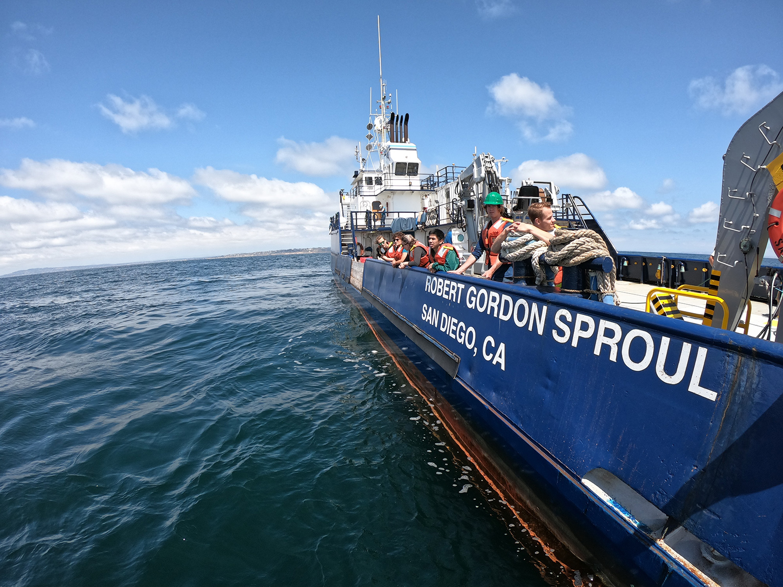 A number of young people stand on the deck of a Coast Guard ship. The foreground is mostly taken up by the royal blue hull of the ship with "Robert Gordon Sproul/San Diego, CA" emblazoned in white.