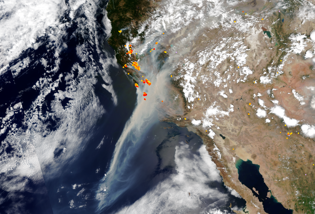 A true-color satellite view of California. Numerous red, orange and yellow dots are sprinkled along the state\'s coastline, indicating fires of varying intensities. Massive amounts of grey smoke obscure the central part of the state and form a plume extending out over the Pacific Ocean.