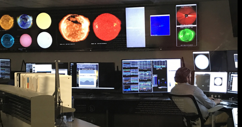 A somewhat dark room filled with desks with multiple computer monitors and a giant monitor with images of the planets at the front of the room. A man observes his computer screen.