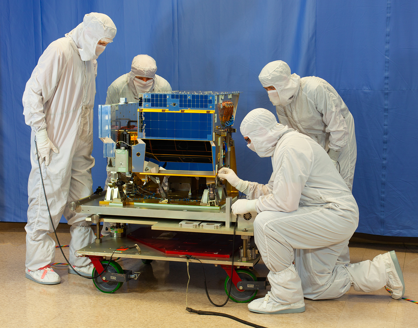 Four technicians in white cleanroom suits, masks and hoods gather around the CrIS instrument on a wheeled cart. The instrument is about the size of a large microwave. The technician in the front on the right kneels and adjusts a small piece of the instrument.