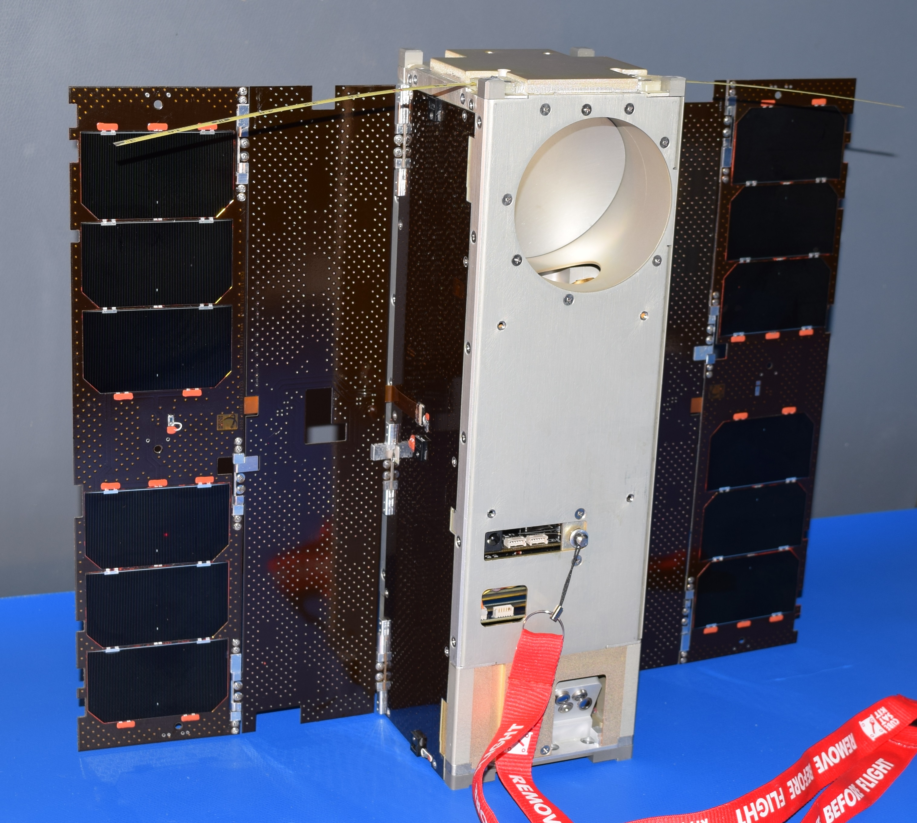 A small satellite, roughly the size and shape of a shoe box, sits on a blue tabletop. To either side of the metal box that forms its base, two long rectangular solar panels are deployed.