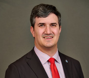 Jordan Gerth with wavy black hair and brown eyes, wearing a black suit, white shirt and red tie