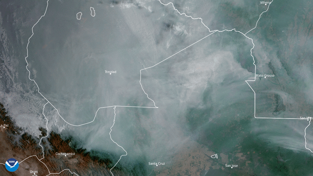 Grayish-brown smoke is seen billowing over brown/green landmass with white clouds above the smoke.