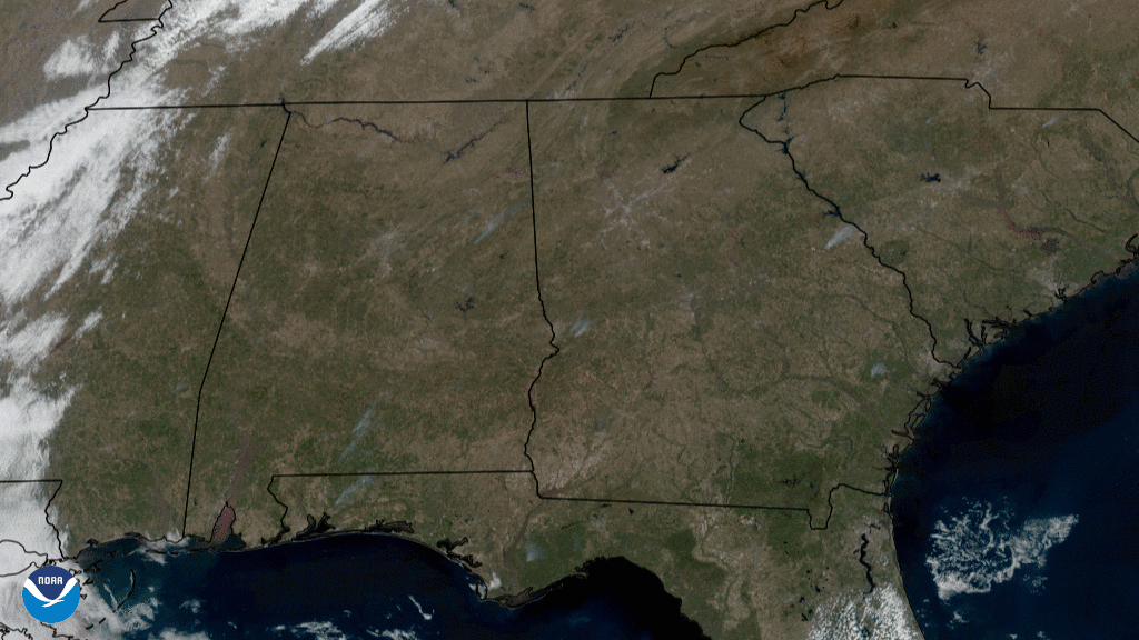 The brown and green landscape of the southeastern U.S. is seen from above with many grayish smoke plumes dotting the landscape and white clouds move above the scene. 