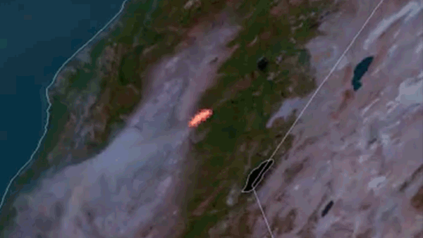 Red fire hot spot with gray smoke emanating from it is seen against the brown/green terrain. 