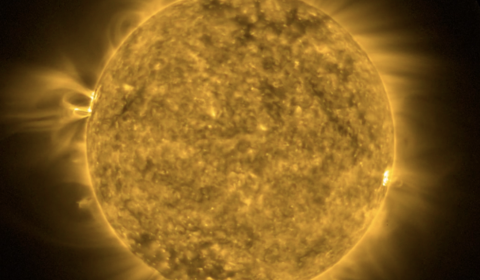 A telescopic image of the Sun zoomed to fill the screen.  Colored a false green to identify the image as taken at a wavelength of 94 Angstroms to show 6 million degree plasma from the solar corona.  Mottled with bright spots and dark regions, a bright spot just right of center stands out.  This spot is the location of a solar flare which heats coronal plasma to millions of degrees revealing the magnetic field loops responsible for the solar flare.