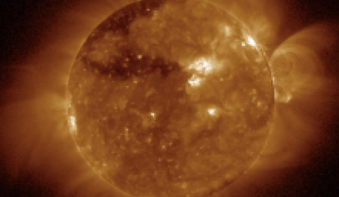 A telescopic image of the Sun, with a disk blocking the direct light from the Sun to reveal the faint, low density, solar corona, or atmosphere, falsely colored red.  Seen stretching radially from the Sun are coronal streamers, areas of higher density.  On the lower right, are arches of erupting magnetic field loops organized into a Coronal Mass Ejection, seen erupting outward from the Sun.