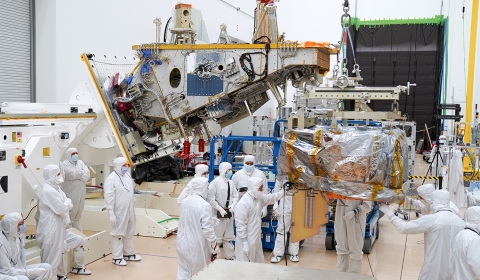 In a clean room, engineers supervise the VIIRS instrument as it is suspended by a lift. The main bus on which it will be fitted onto is nearby.