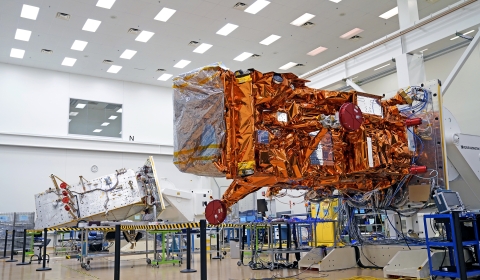 JPSS-3 is visible in the foreground of the photo as a large rectangle covered in orange mylar with hardware attached.  JPSS-4 is in the background of the photo, in the bottom left.  It appears as a silver rectangle with wires and hardware attached.  Both spacecraft are attached to white rollover fixtures.  The satellites sit in a large, white room with rows of lights on the ceiling and a large observation window in the top, center of the photo.  The room has a tan floor with black stanchions and black and y