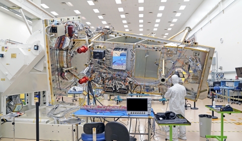 The JPSS-4 spacecraft bus appears in the center of the image as a silver rectangle with wires and hardware covering the surface while a person in a white, bunny suit works towards the right-hand end of the spacecraft. The satellite is attached to a white, rotating fixture in a white room with florescent lights lining the ceiling. A blue desk with two chairs and a monitor sits in the bottom center of the image. Two green, rolling tables also appear in the bottom of the image.