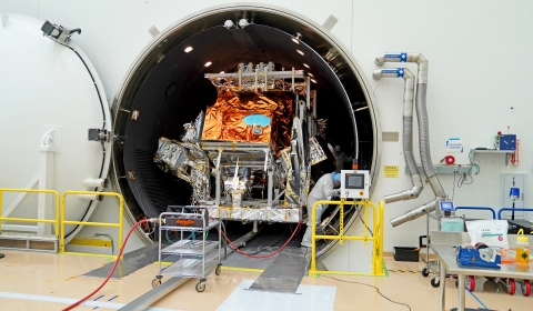 A large, tubular champer sits open in a large, white clean room with its white door seen on the left against a white walll. The silver and orange mylar-covered JPSS-3 satellite sits inside the chamber while a person in a white bunny suit works on the right side of the spacecraft. Outside of the chamber there is yellow railing on both sides of the tube. There are silver pipes attached to the wall next to the chamber opening. Outside of the chamber, there is a tan and white floor, silver carts and tables wit