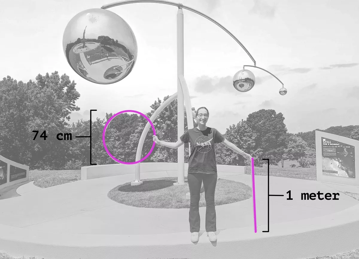 Photo of a person holding a hoop and yard stick in front of an outdoor sculpture.