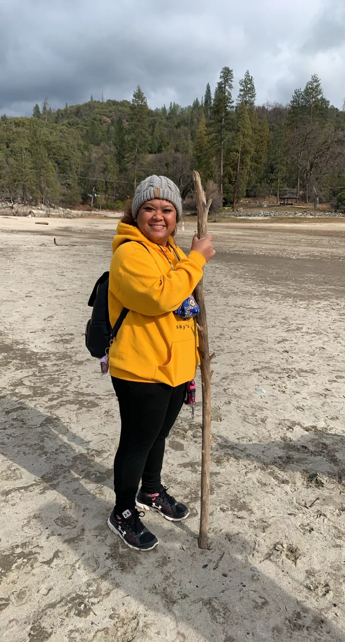 Larafae Santos, seen here hiking on a sandy area with conifer trees in the background. She is dressed in a yellow sweatshirt, black pants, a gray hat, backpack, and is holding a walking stick. 