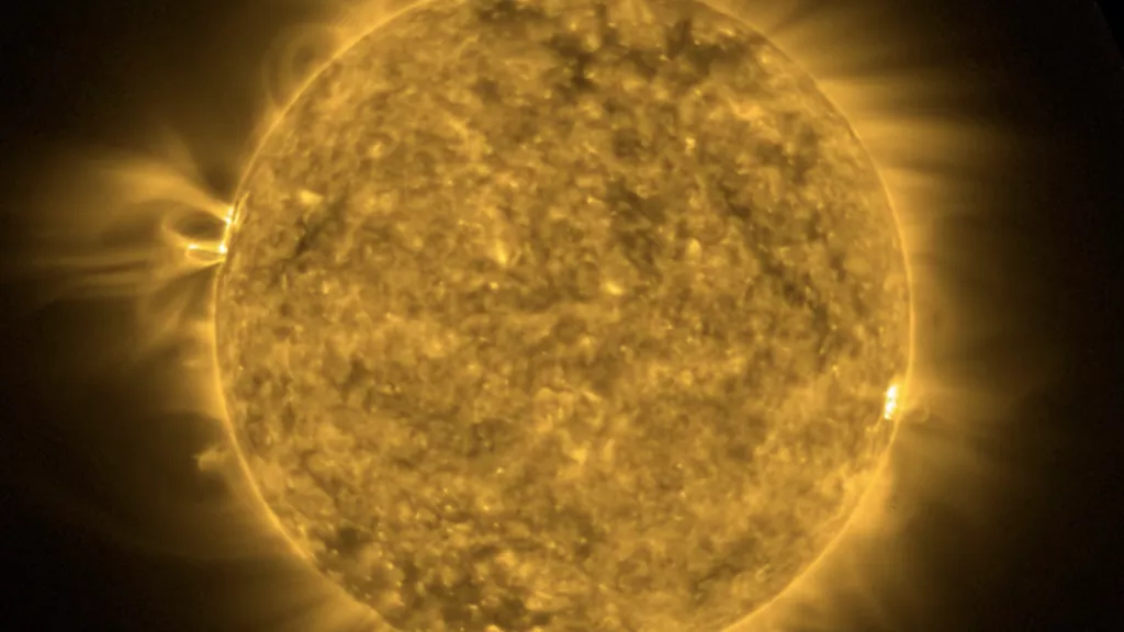 A telescopic image of the Sun zoomed to fill the screen.  Colored a false green to identify the image as taken at a wavelength of 94 Angstroms to show 6 million degree plasma from the solar corona.  Mottled with bright spots and dark regions, a bright spot just right of center stands out.  This spot is the location of a solar flare which heats coronal plasma to millions of degrees revealing the magnetic field loops responsible for the solar flare.