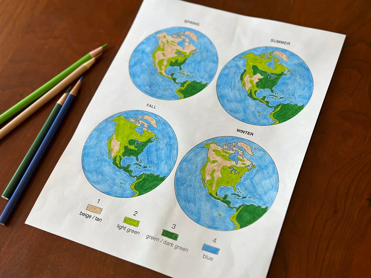 Paper with colored in maps of the Earth on a wooden tabletop, with colored pencils.