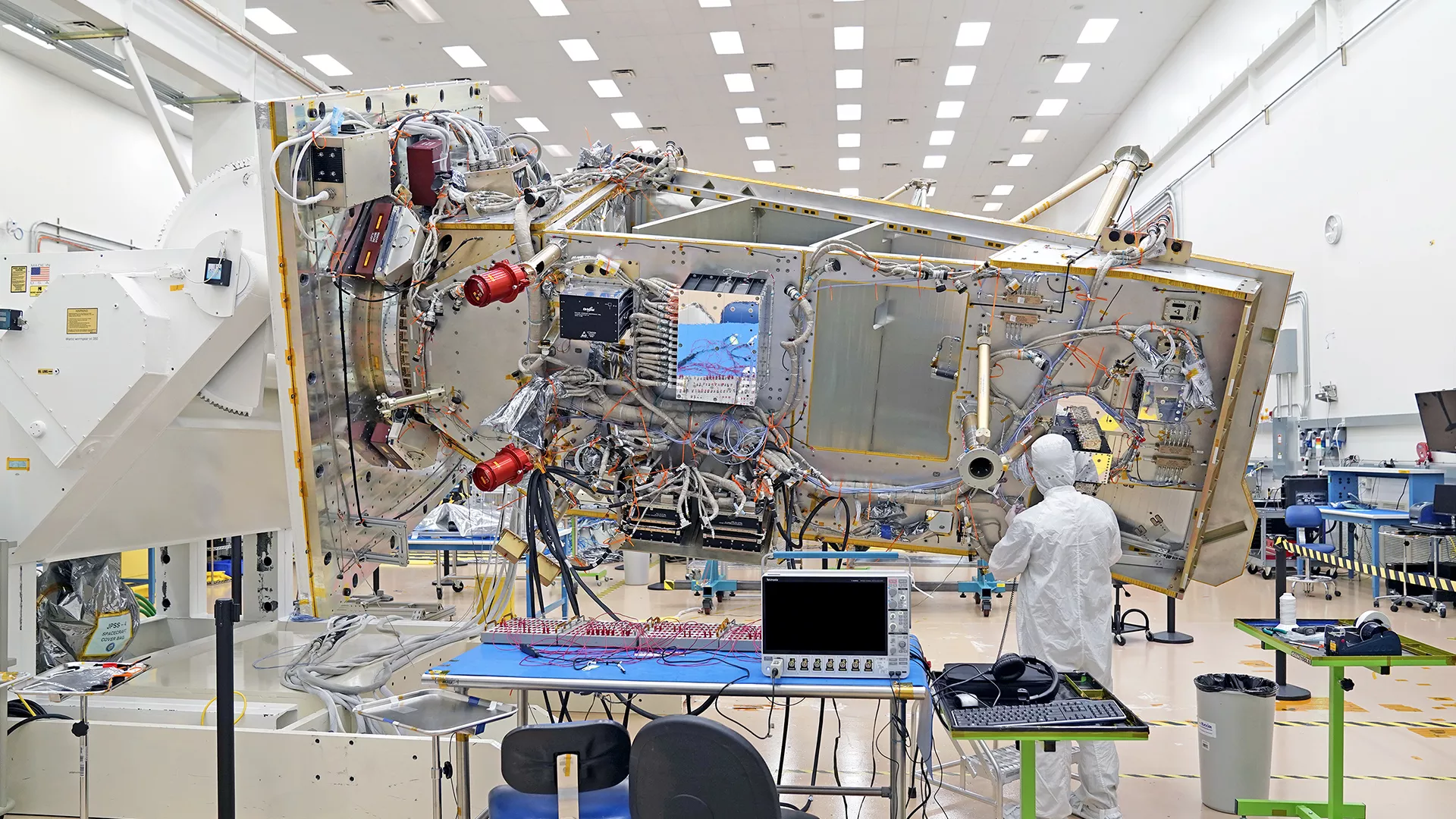 The JPSS-4 spacecraft bus appears in the center of the image as a silver rectangle with wires and hardware covering the surface while a person in a white, bunny suit works towards the right-hand end of the spacecraft. The satellite is attached to a white, rotating fixture in a white room with florescent lights lining the ceiling. A blue desk with two chairs and a monitor sits in the bottom center of the image. Two green, rolling tables also appear in the bottom of the image.