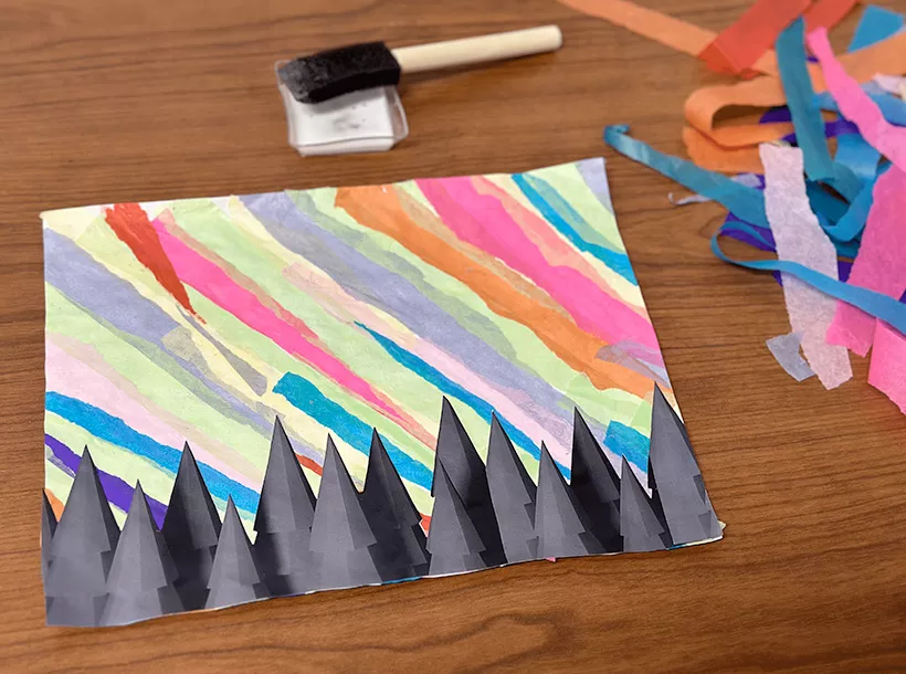 A photo of an completed art project on a desk top, along with strips of tissue paper and a brush and glue. Artwork is a paper with colorful streaks and black paper cut-outs resembling a coniferous tree tops.