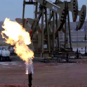 The image depicts an oil field at dusk, characterized by a large, controlled burn-off flame from a gas flare. In the background, an oil pump jack is silhouetted against the fading light of the day. The sky is a muted blue and the ground is bare, brown soil. Storage tanks and additional infrastructure are visible in the midground.
