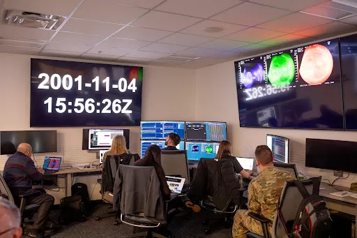 People at the space weather prediction center viewing a television screen.