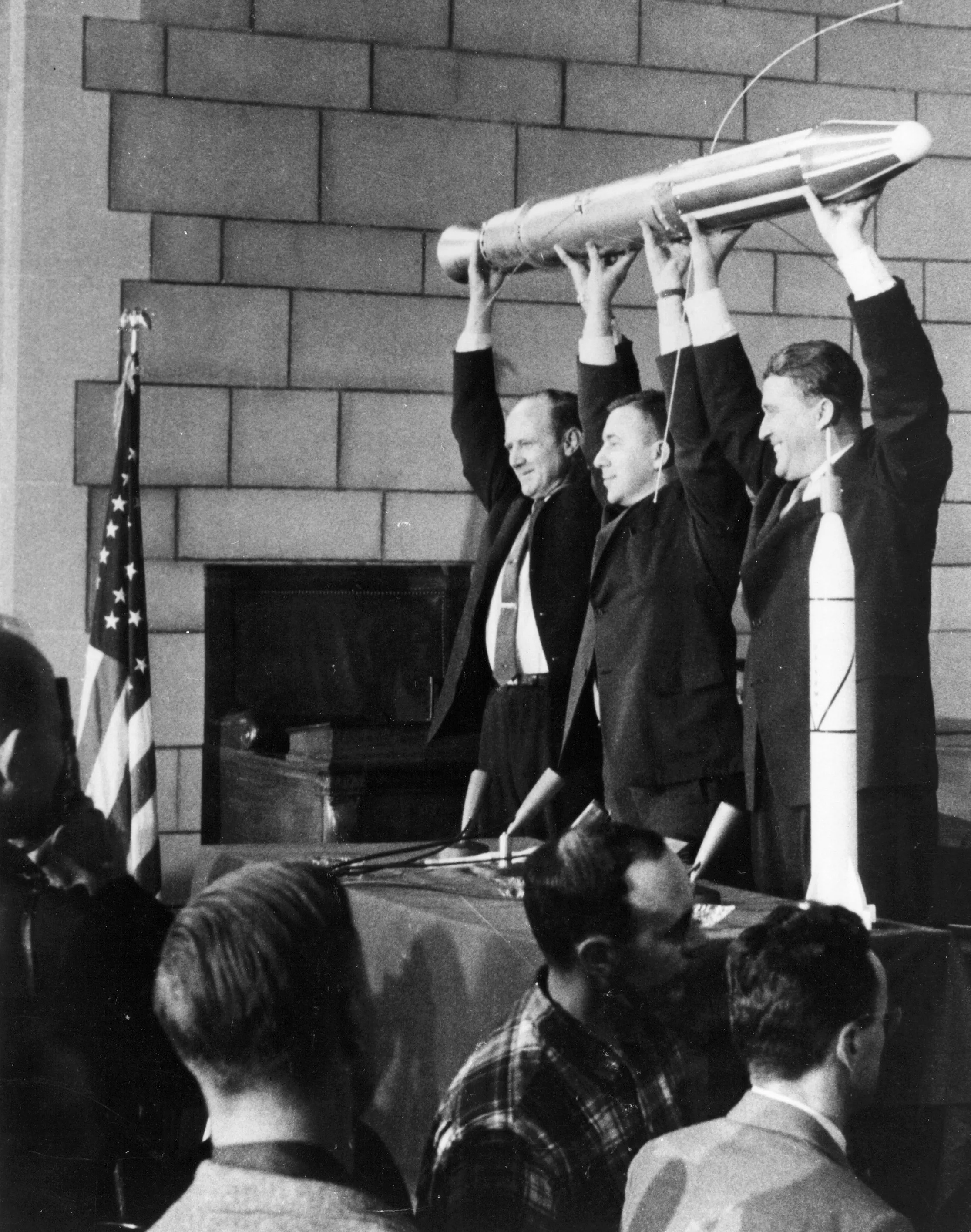 Three men in front of an audience in a black-and-white photo from 1958, dressed in suits and holding up a small rocket. 