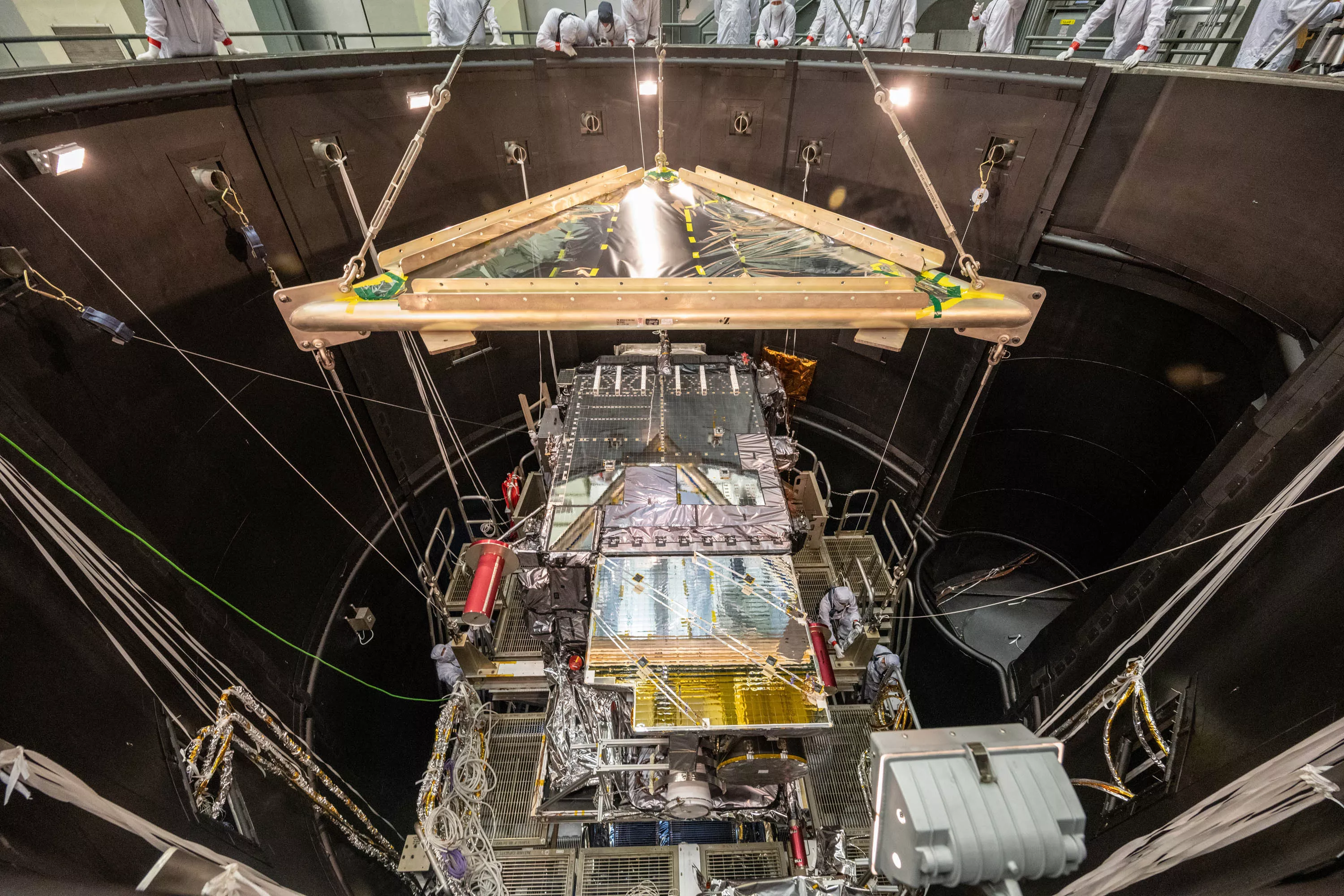 The satellite is seen near the top of a very large chamber with many cables lowering it into the chamber.  Technicians in clean suits look on. 