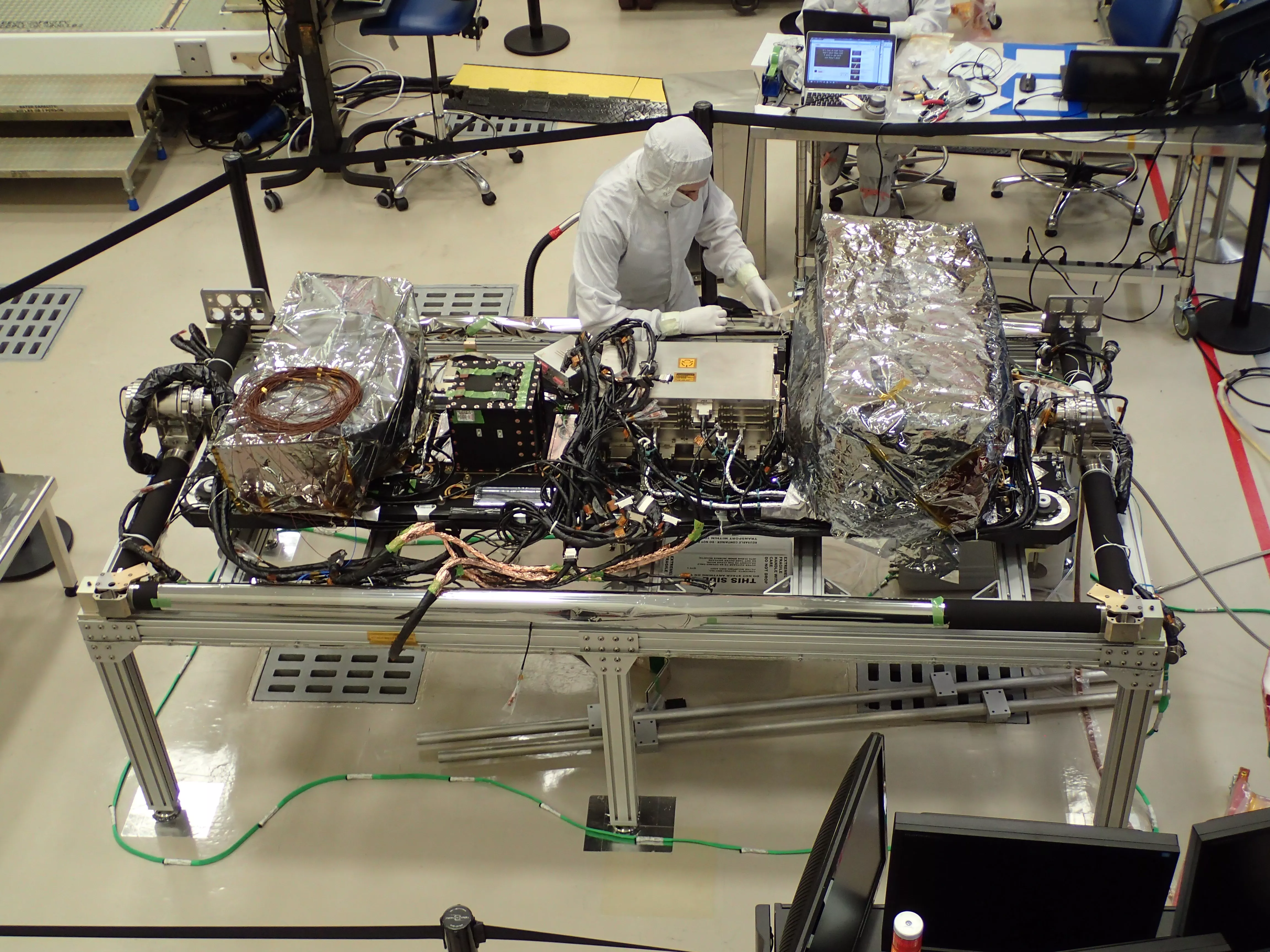Photo of a clean room and two satellite instruments laid on their sides, with suited workers all around its perimeter.