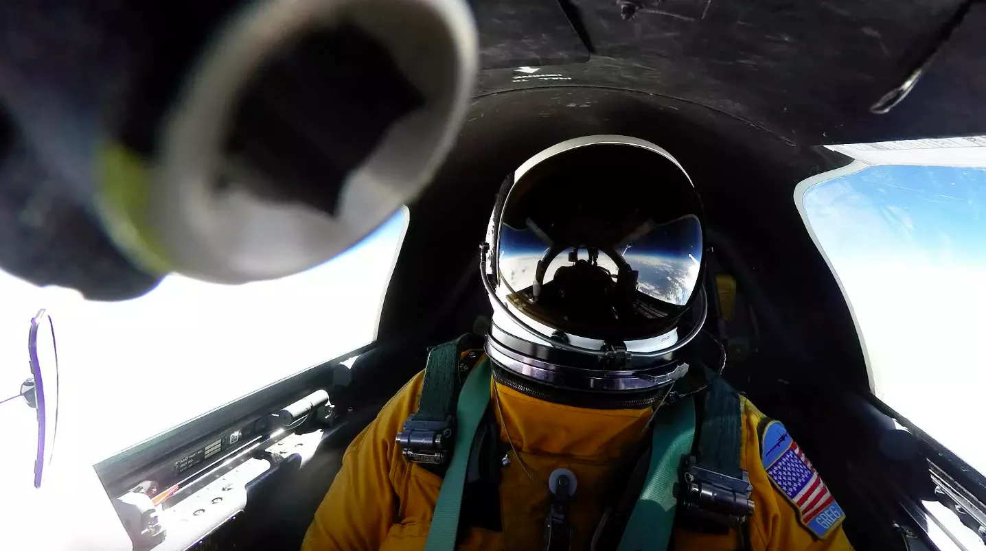Pilot, in orange jumpsuit and helmet, sitting in airplane cockpit.