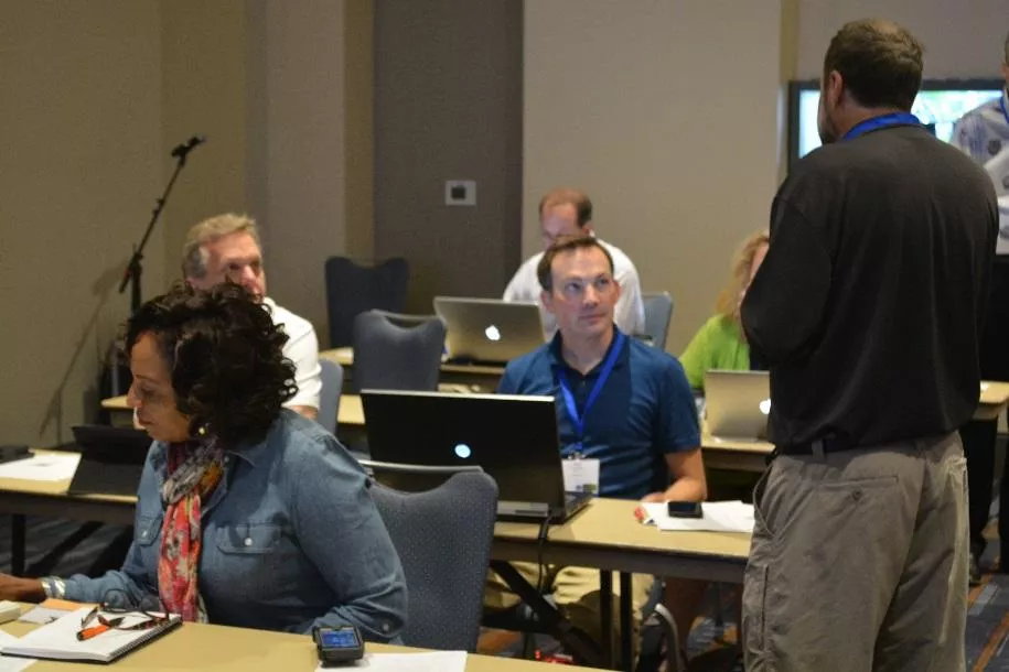 People seated at rows of desks with laptops in front of them converse with course instructor.